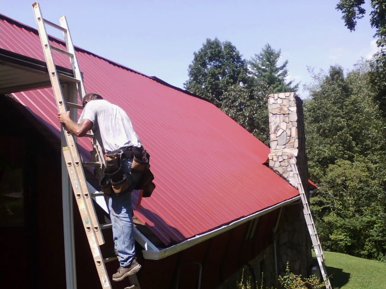 Roofer on a ladder beside a red metal roof and stone chimney — Soffit & Fascia Repair by Roofing Brothers in Wichita Falls
